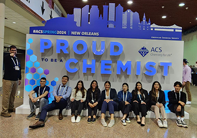 A group representing UDM's Chemistry Club poses for a photo in front of a display for the ACS New Orleans 2024 conference. The display has text that reads "proud to be a chemist"