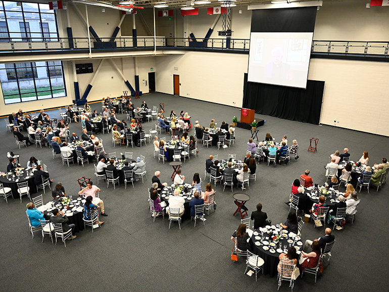 More than 100 people sit around tables inside of the Student Fitness Center, watching a video being player on a projector screen.