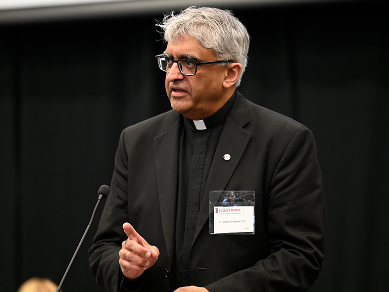 A man wearing a nametag that reads Fr. Gilbert Sunghera, S.J. speaks from a podium indoors.