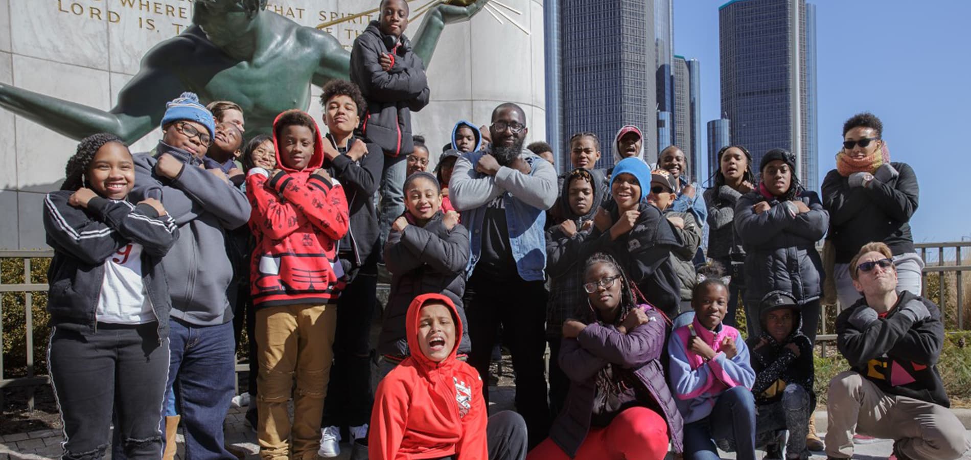 Michael Ford stands outdoors in front of an iconic statue in downtown Detroit, surrounded by more than a dozen children in sweatshirts and coats.