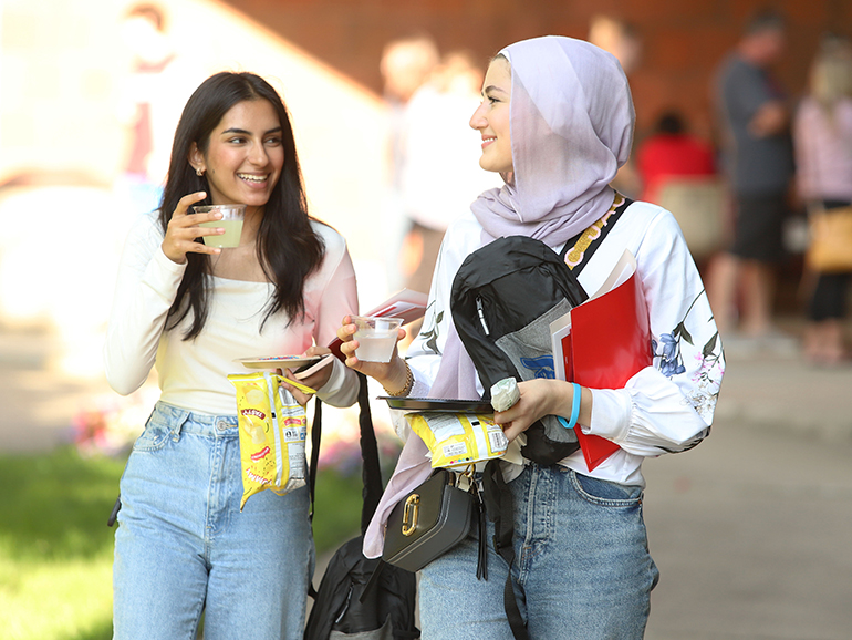 Two students carrying drinks and other belongings outdoors smile as they talk to one another on the McNichols Campus.