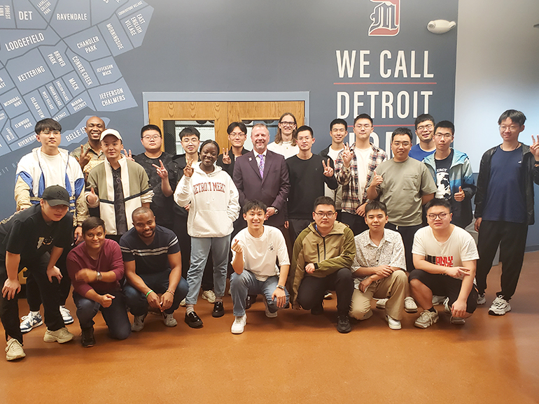 Nearly two dozen students and President Taylor stand inside of the Engineering Building, with a We Call Detroit Home mantra on the wall behind them.