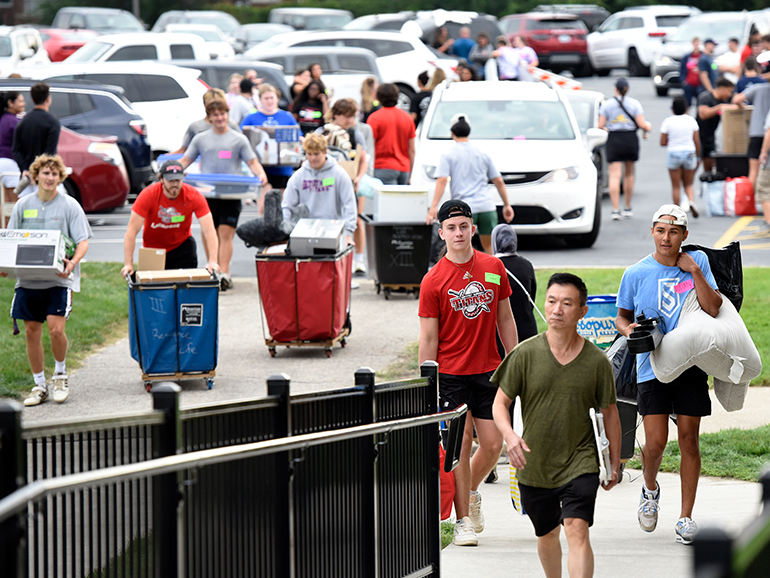 Students walk, carry and push carts outdoors on the sidewalk towards Shiple Hall, with many other people and cars in the background.