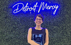 Megan Novell holds an award while standing in front of a green backdrop with a blue, neon sign that spells Detroit Mercy.