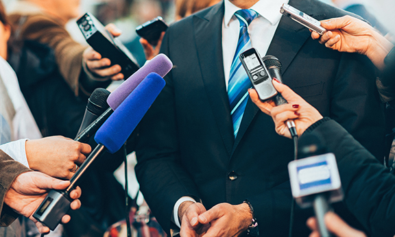 Shows man in a suit with media microphones pointed at him