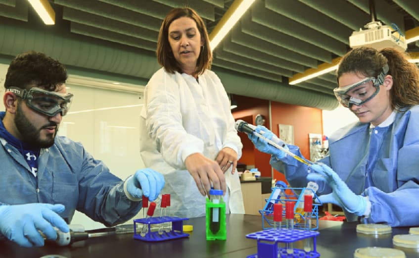 bio prof showing two undergrad gloved and smocked bio students a vial