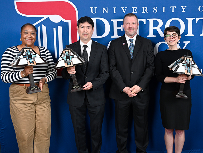 Four people, three holding lamps, stand and pose for a photo inside with a blue University of Detroit Mercy backdrop behind them.