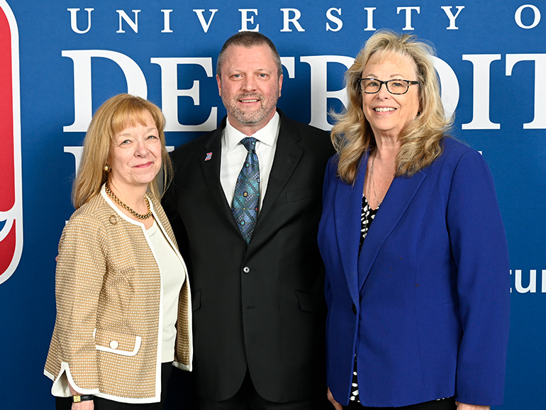 Three people stand smiling in front of a blue University of Detroit Mercy backdrop.