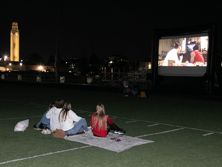 A group of students sit on the Titan Field turf for the screening of Back to the Future. In the background, the clocktower is visible and lit up.