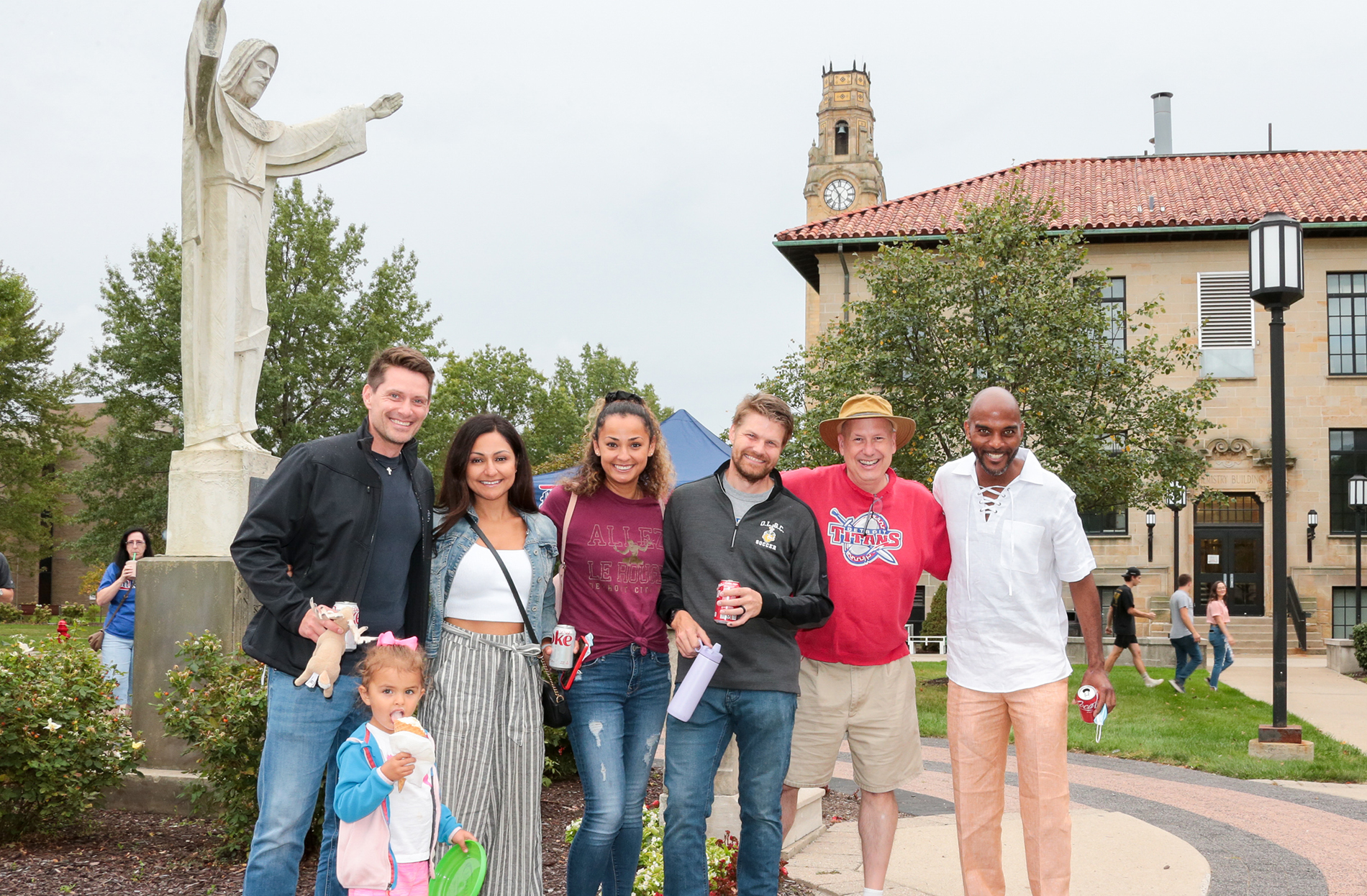 A group of adults and one child pose for a photo in front of Detroit Mercy's Jesus statue. In the background, the clocktower, buildings and other people are visible.