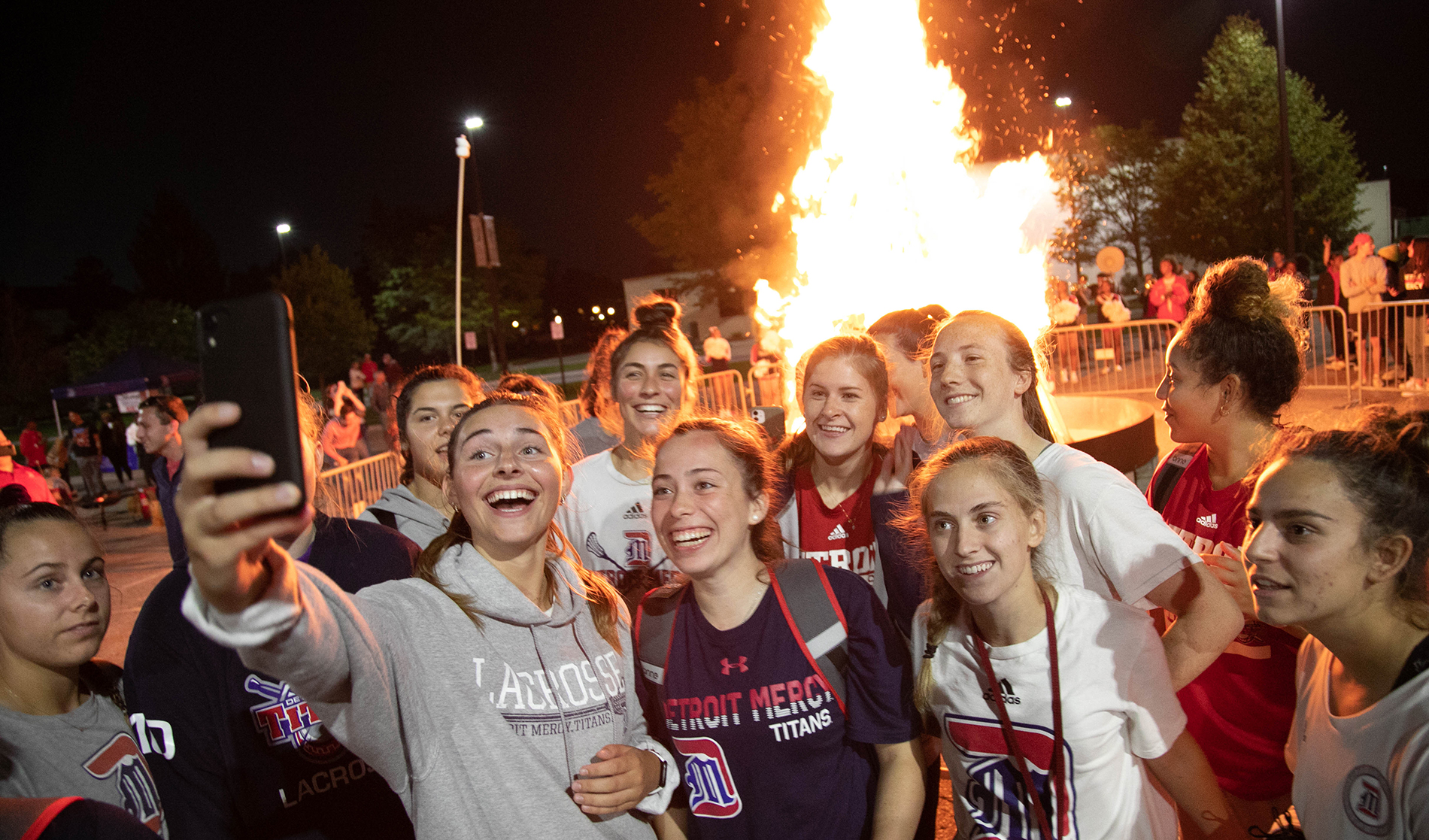 A group of female students wearing Detroit Mercy lacrosse gear take a selfie next to the Titan Tailgate bonfire.