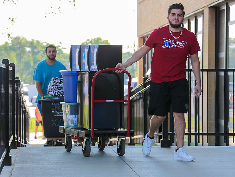 Two students, one wearing a University of Detroit Mercy shirt, wheel belongings into Shiple Hall during move-in day.