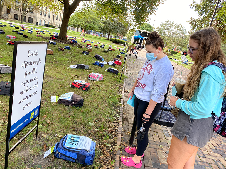 Two students look at a message on one of the backpacks in the Send Silence Packing exhibit. A sign next to the backpack reads “Suicide affects people from ALL ages, races, genders and socioeconomic groups.”