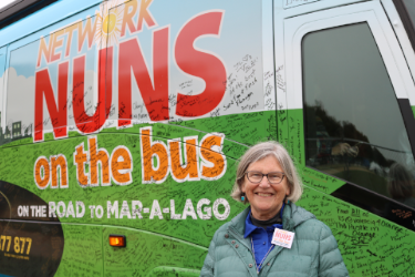 A photograph of Sr. Simone Campbell, SSS, in front of the Network Nuns on the Bus vehicle. The text on the bus includes "on the road to Mar-a-Lago" and many signatures on the bus are visible.