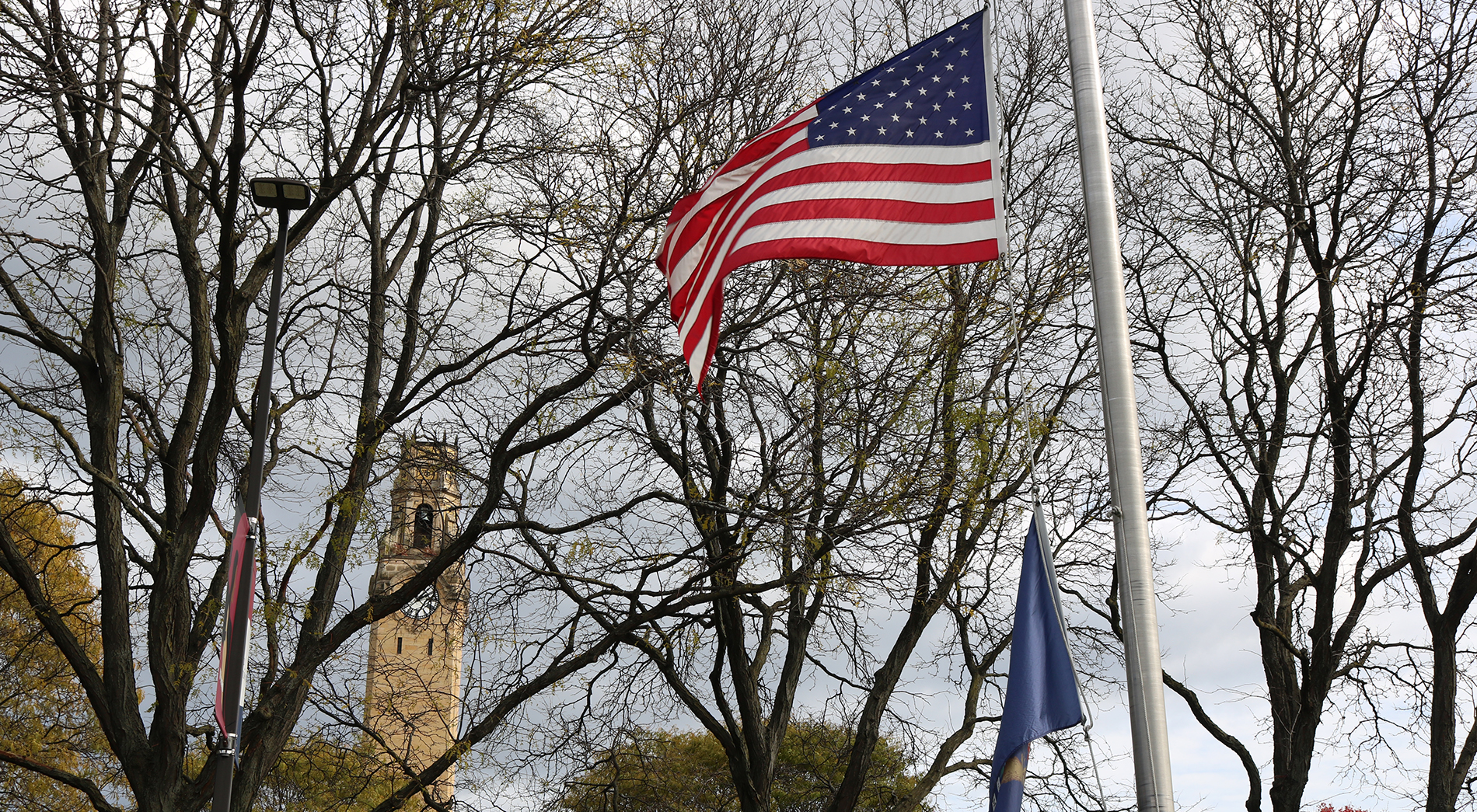An American flag waves in the breeze on Detroit Mercy's McNichols Campus. In the background, the clocktower is visible, beneath trees and foliage.