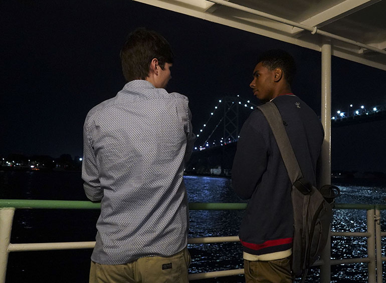 Two students talk while standing against the railing of a boat during the boat cruise. The Ambassador Bridge's lights are visible in the background.