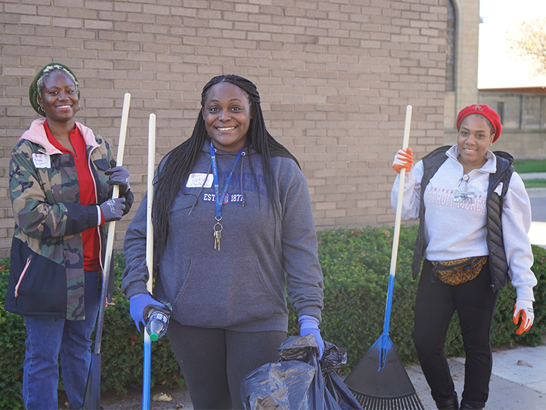 Three women in Detroit Mercy gear stand outside with rakes and pose for a photo.