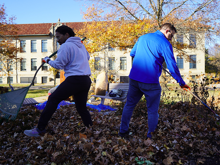 Two men rake leaves outdoors on the McNichols Campus with buildings in the background.