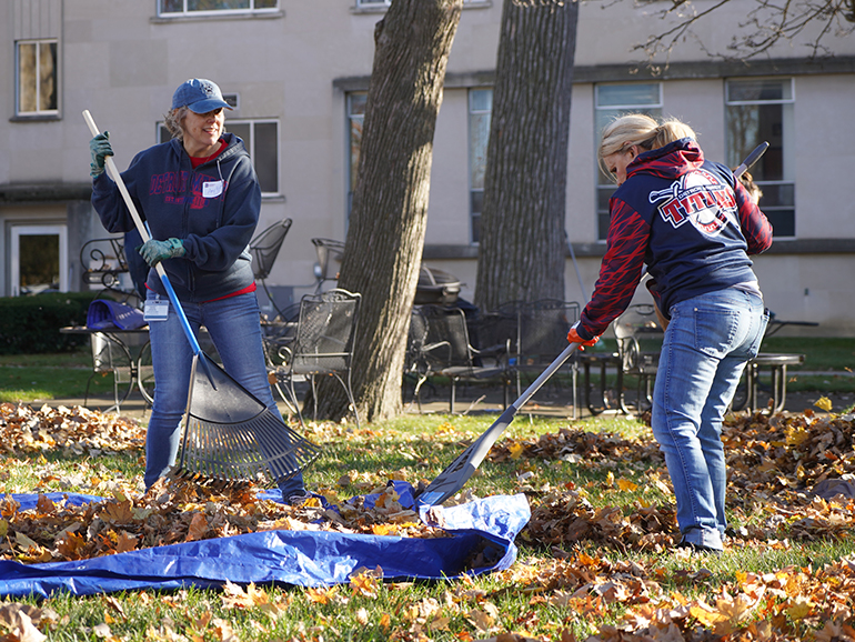 Two women wearing Detroit Mercy attire rakes leaves with Lansing-Reilly Hall in the background on the McNichols Campus.