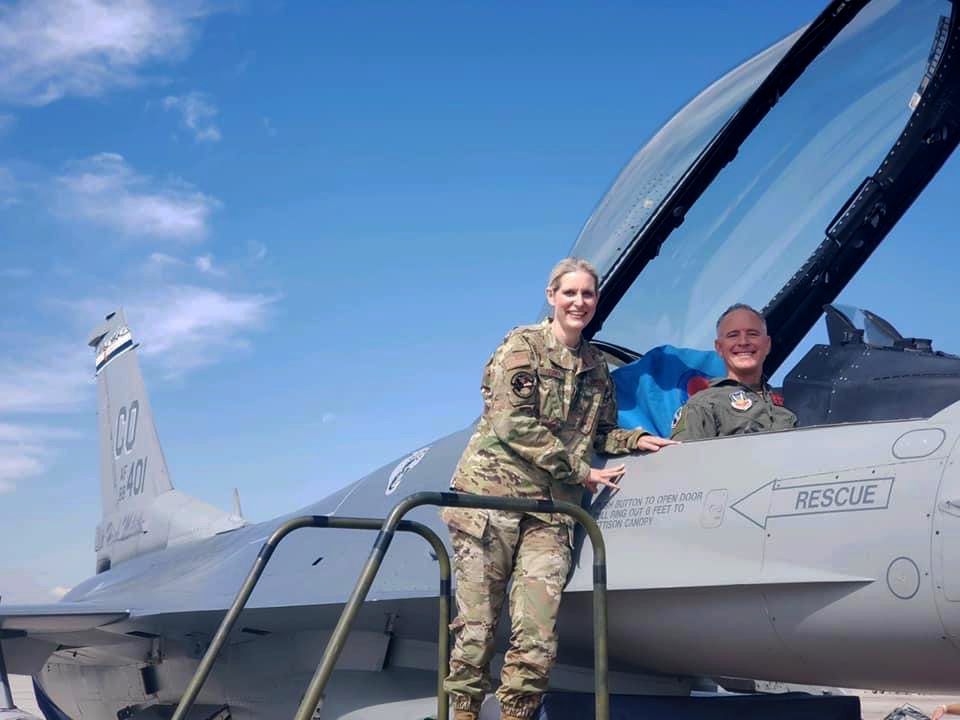 One person stands on a ladder and leans on a fighter jet while posing for a photo outdoors.