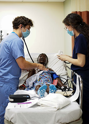 Two students wearing scrubs perform a healthcare screening on a simulation mannequin.