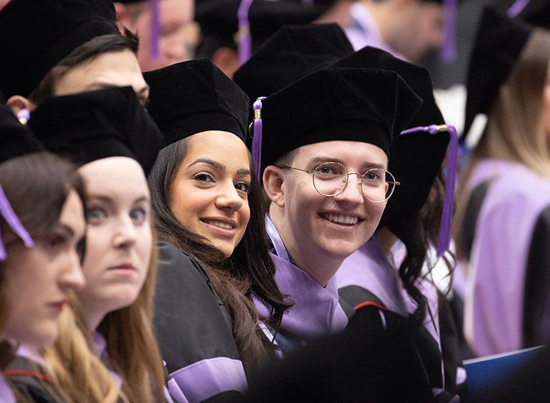 In a crowd of graduates, two smile while looking at something during commencement.