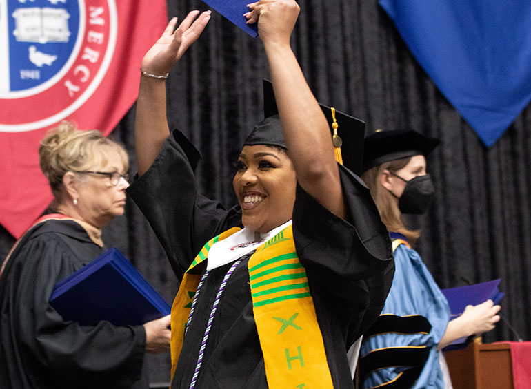 A graduate lifts up her arms in celebration while walking across the stage during commencement.