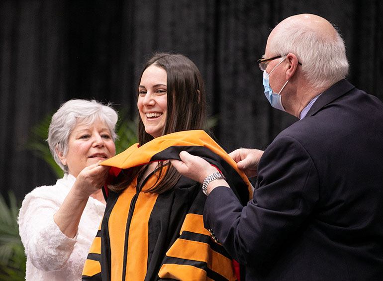 A graduate smiles as she receives her hood from two people.