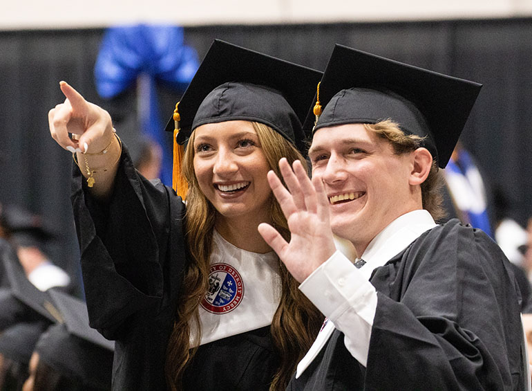 A graduate points to someone during commencement while the graduate next to her waves in that area.