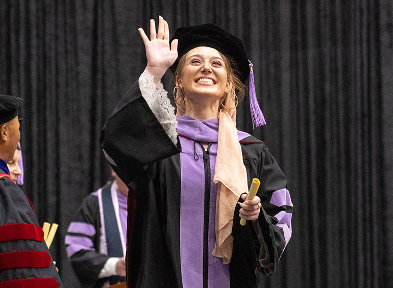 A graduate smiles and waves to the crowd after walking across the commencement stage.