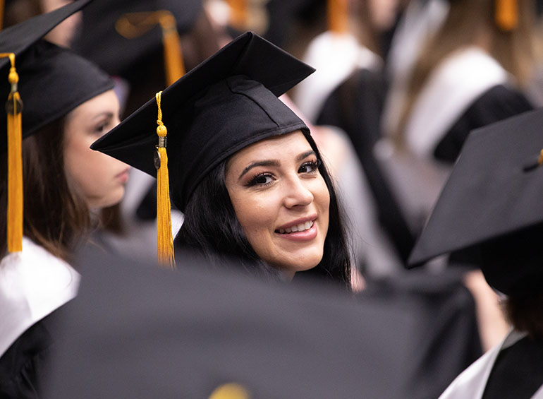 A graduates smiles for a photo while sitting during commencement. Other caps and tassels of graduates surround her face.