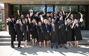 A group of students happily raise their degrees in the air as they pose for a photo outside of McNichols Library.
