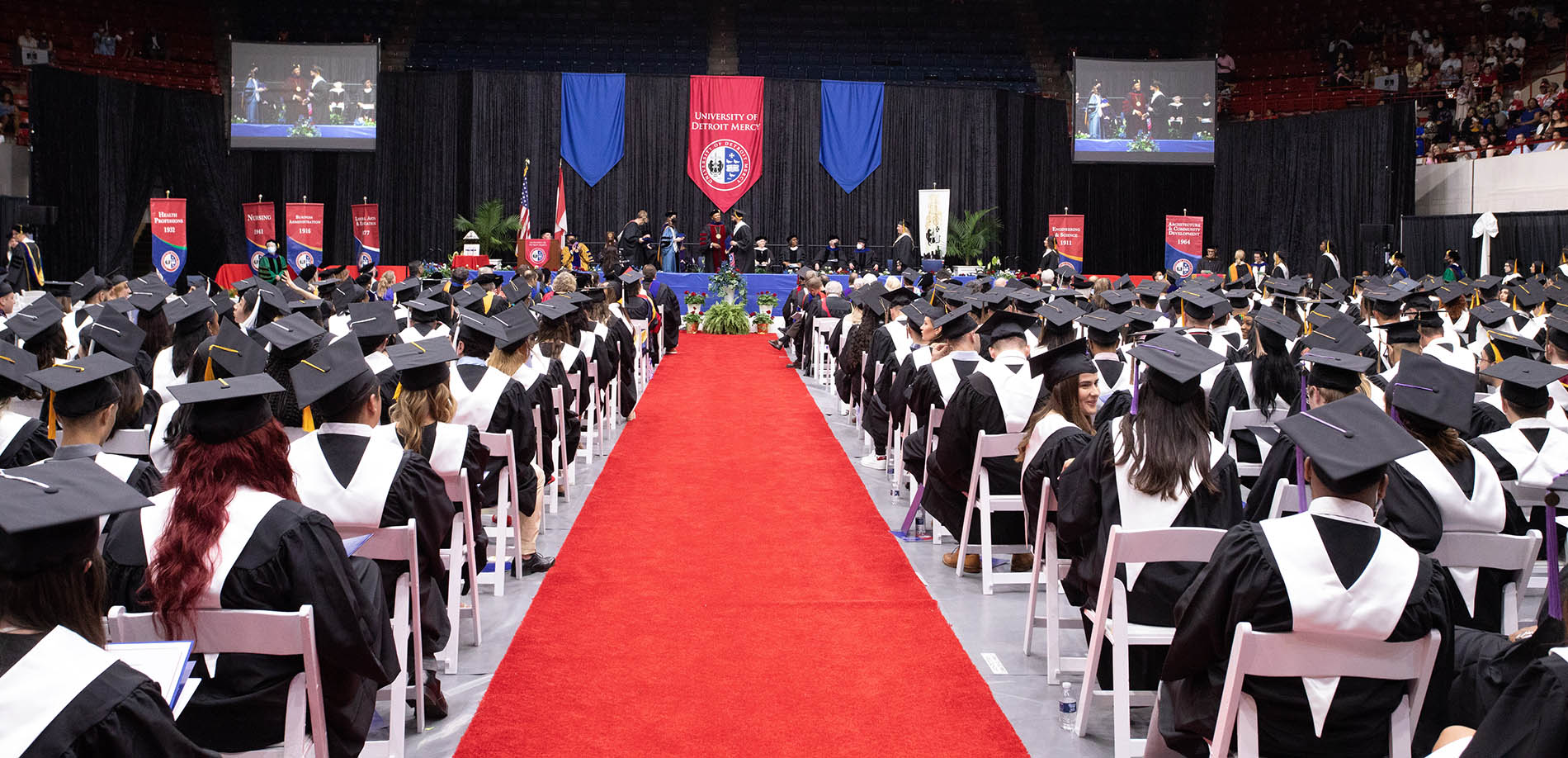 The graduates patiently wait in their seats while the other graduates receive their degrees on stage in Calihan Hall. A big red banner on stage reads, “University of Detroit Mercy.”
