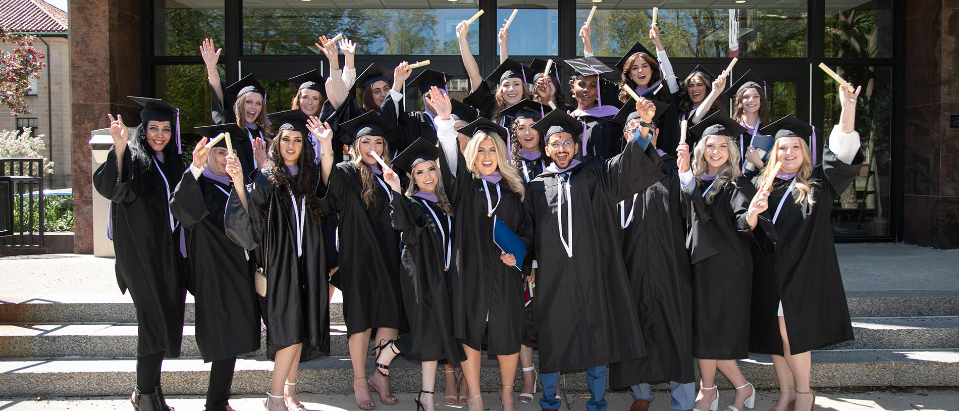 A group of students happily raise their degrees in the air as they pose for a photo outside of McNichols Library.