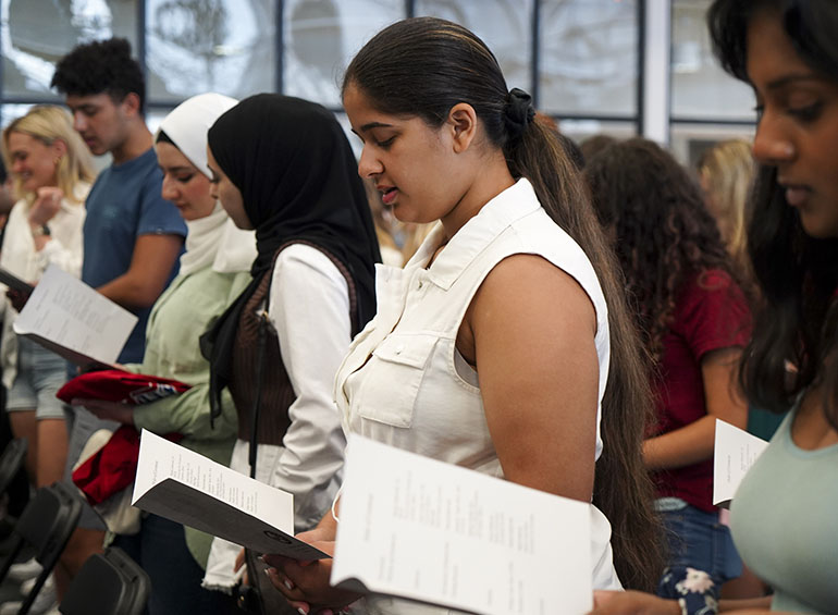 A student participates in first-year convocation by reading their program. Other students who are also reading are visible in the background and foreground. 
