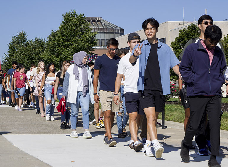 A student flashes the peace sign as students walk in a line down Kassab Mall to start first-year convocation.