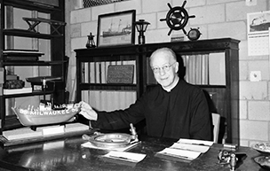 Father Edward Dowling sits inside at a desk with his hand holding a model Great Lakes ship.