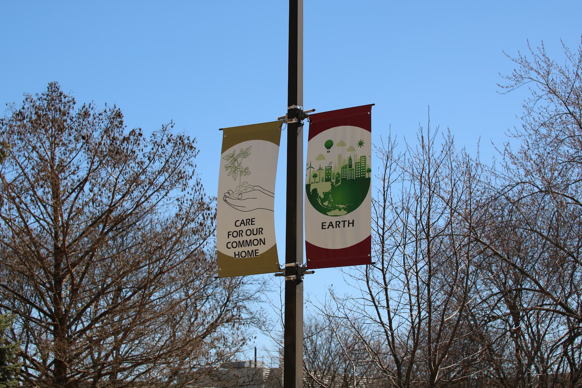 An outdoors photo on the McNichols Campus shows to signs, reading 'Care for Our Common Home' and 'Earth' with trees in the background.