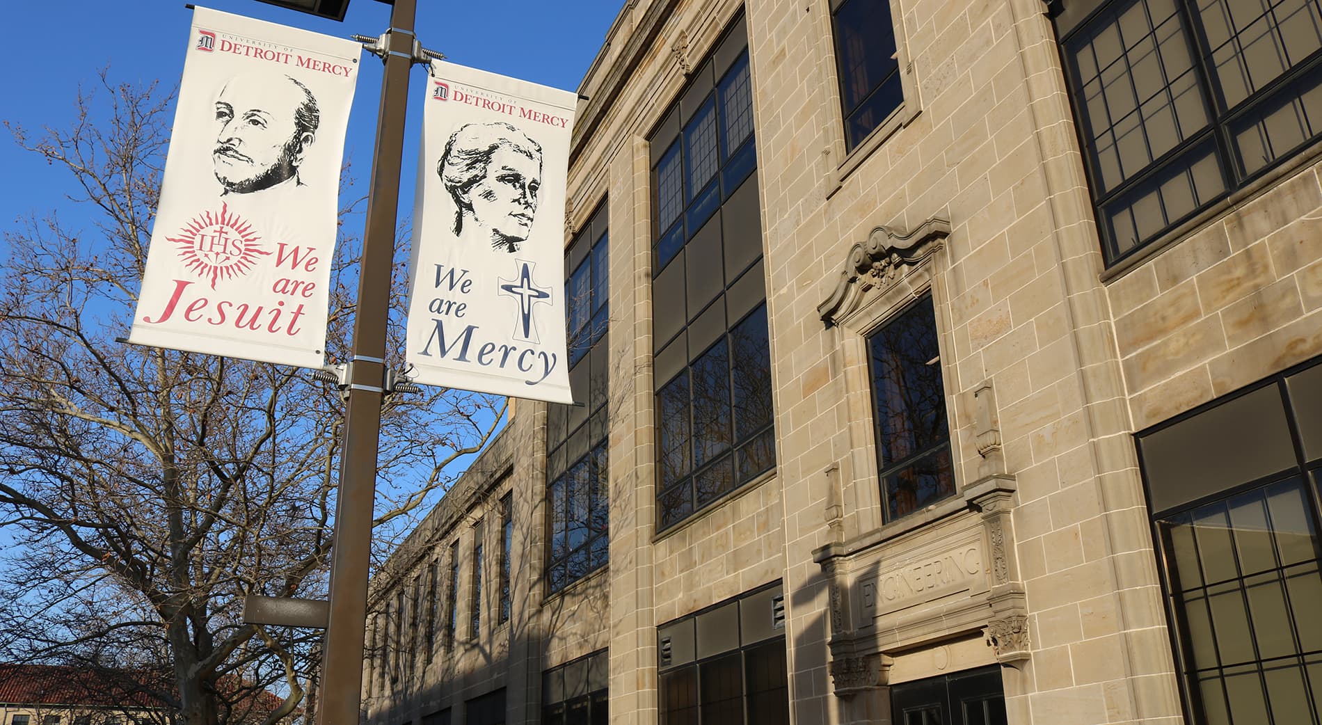 An exterior photo of the Engineering Building's side entrance, with We are Mercy and We are Jesuit light pole banners visible.