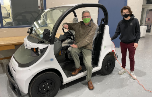 Professor Alan Hoback sits in an electric vehicle, while student Brendan Lubiarz stands near the rear of the vehicle, for a posed photograph.