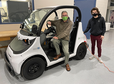 Professor Alan Hoback sits in an electric vehicle, while student Brendan Lubiarz stands near the rear of the vehicle, for a posed photograph.