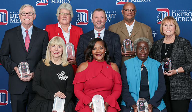 Eight people pose for a photo, three sitting and five standing, with a blue University of Detroit Mercy banner behind them.
