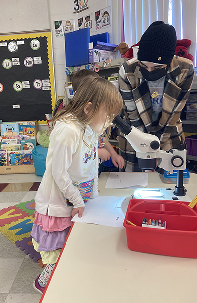 A kindergartener looks into a microscope as a Detroit Mercy student assists.