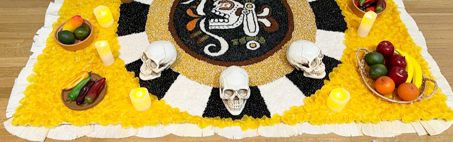 An ofrenda is shown with bowls of plastic fruit, a plate of plastic peppers, fake candles and skulls sit on top an yellow, white, black and tan ornamental sheet indoors.