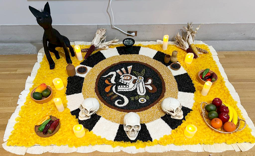 An ofrenda is shown with bowls of plastic fruit, a plate of plastic peppers, fake candles and skulls sit on top an yellow, white, black and tan ornamental sheet indoors.
