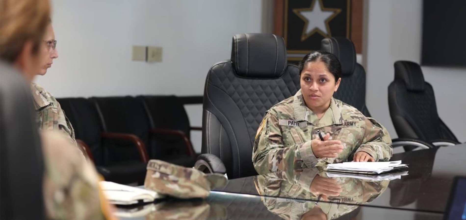 Tanvi Patel in camouflage Army attire sits in a conference room, with two other people.