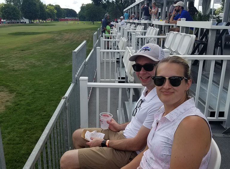 Two people sit in chairs on a raised platform outdoors during the Rocket Mortgage Classic.