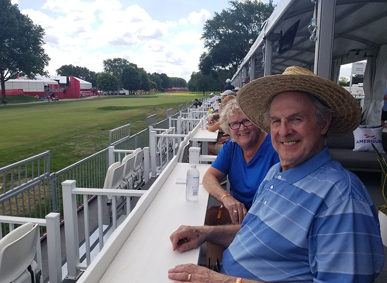 Two people sit in chairs on a raised platform outdoors during the Rocket Mortgage Classic. 