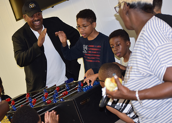 Multiple people cheer while standing and playing around a foosball table indoors.