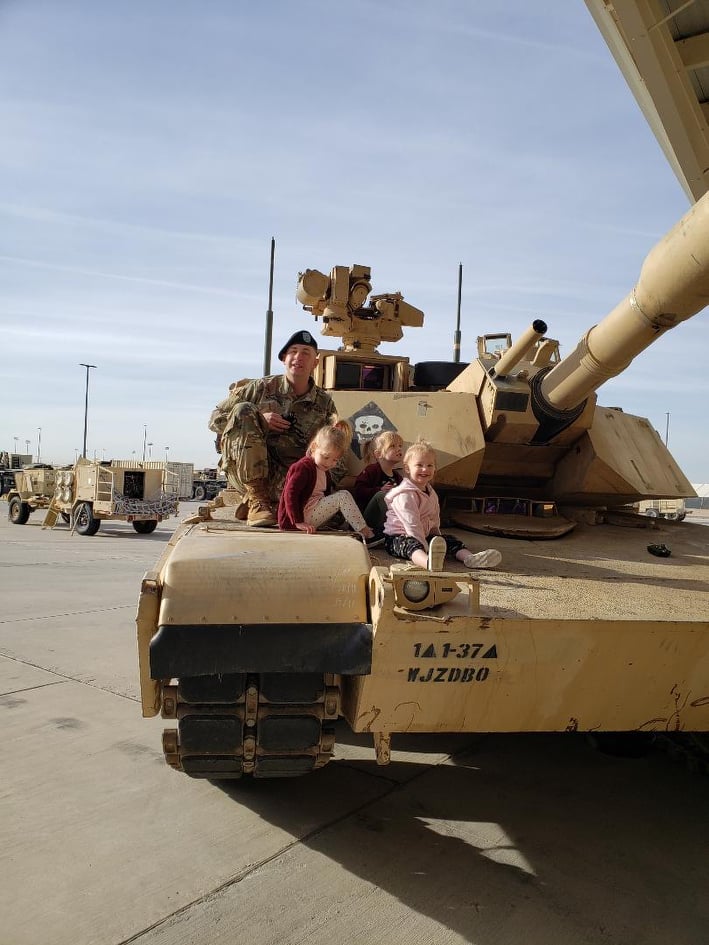 Three people sit on top of an armored vehicle while posing for a photo outdoors.